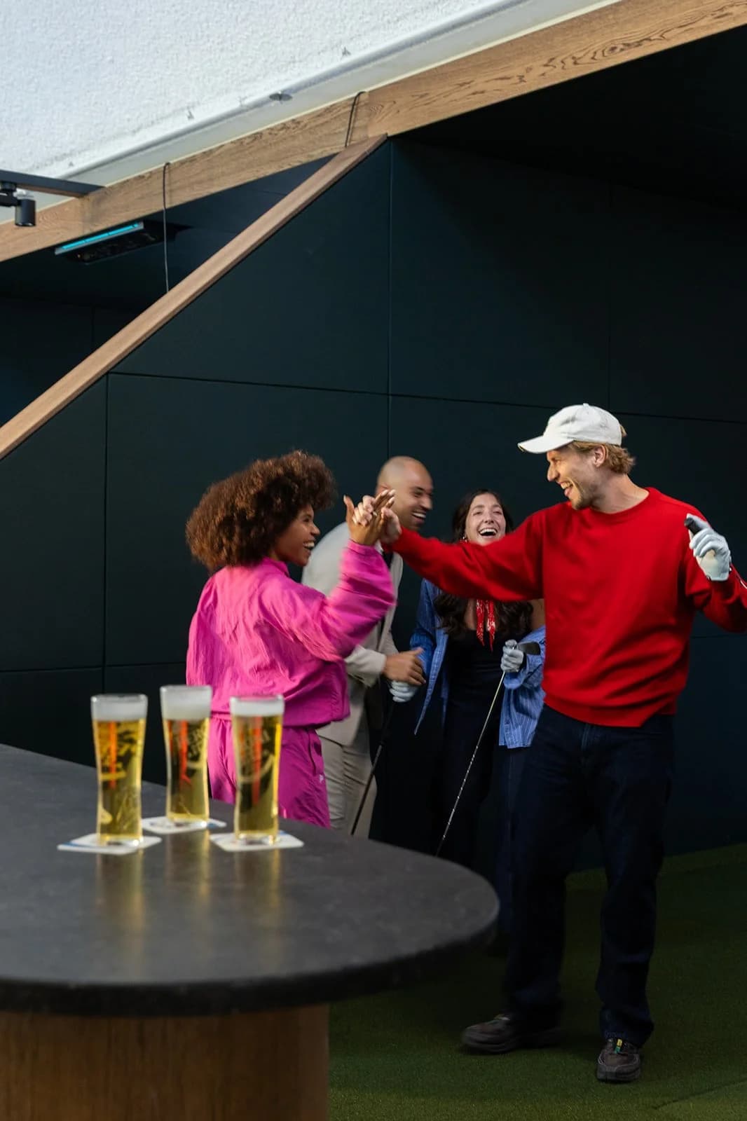 Vieren vier vrienden die lachen en elkaar high-fiven tijdens het spelen van golf in een indoorsportfaciliteit met vier glazen bier op de tafel.